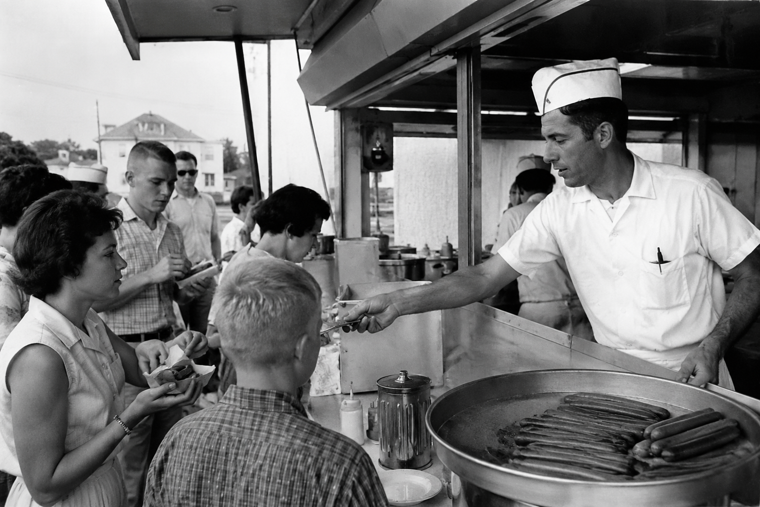 Vintage black and white photo of a 1960s hot dog stand counter with a server in a paper cap
